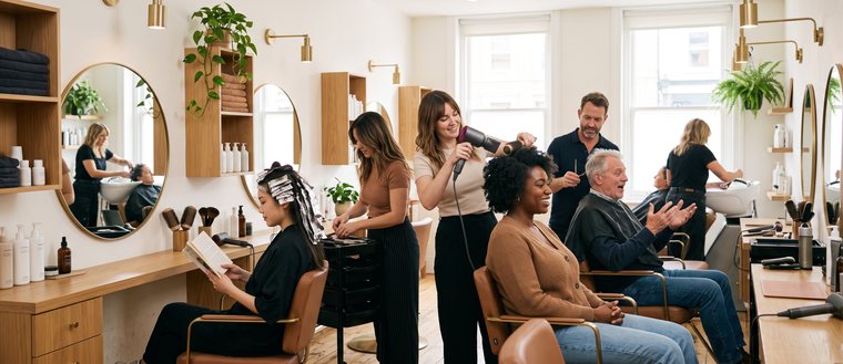 Heller Friseursalon im Betrieb: an drei Plätzen werden Kundinnen und Kunden gefönt, geschnitten und gefärbt, im Hintergrund das Waschbecken